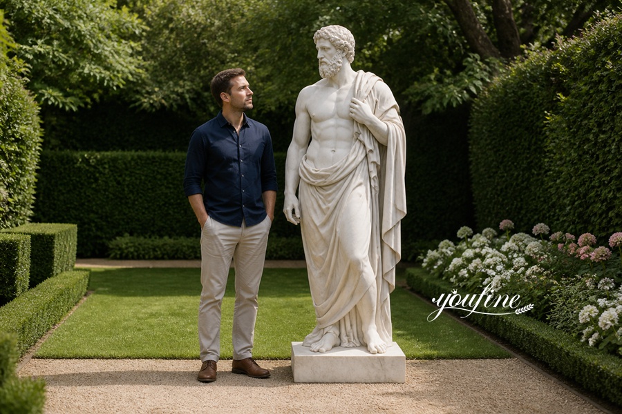 A man in a blue shirt and beige trousers standing beside an intricately carved white marble statue in a manicured garden.
