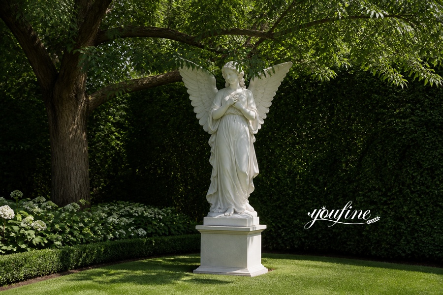 White marble angel sculpture with wings standing on a pedestal in a lush green garden, surrounded by trees and bushes.