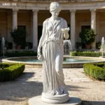 Elegant white marble statue of a woman holding a bowl and pitcher, placed near a circular fountain surrounded by hedges and classical columns.