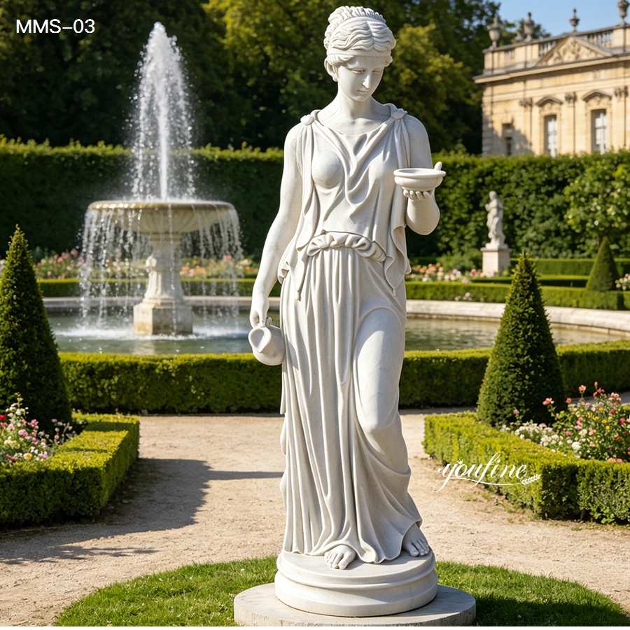 Graceful marble goddess statue holding a bowl in one hand, displayed in a manicured garden with a fountain and greenery in the background.