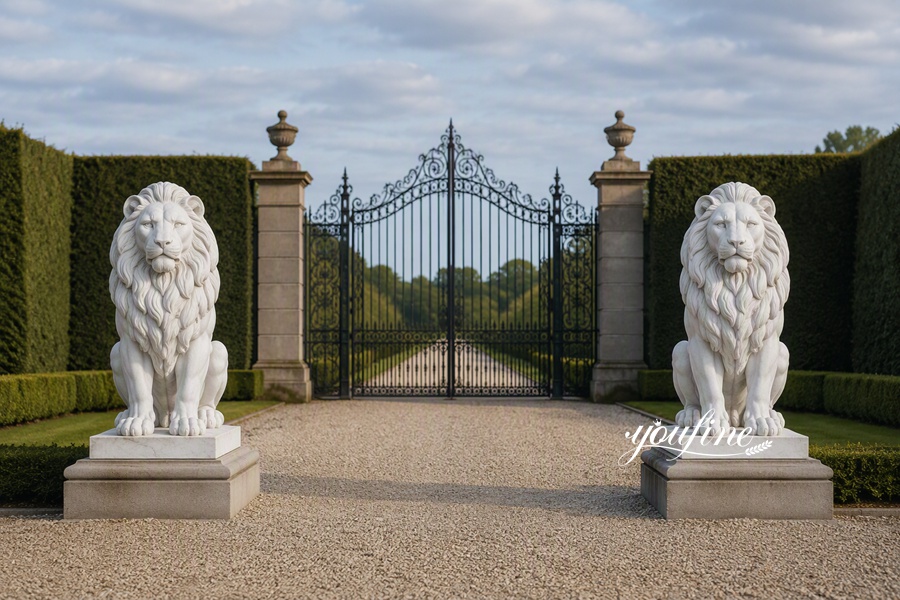 Two intricately carved white marble lion statues placed on pedestals symmetrically flank a grand wrought iron garden gate in a landscaped setting.