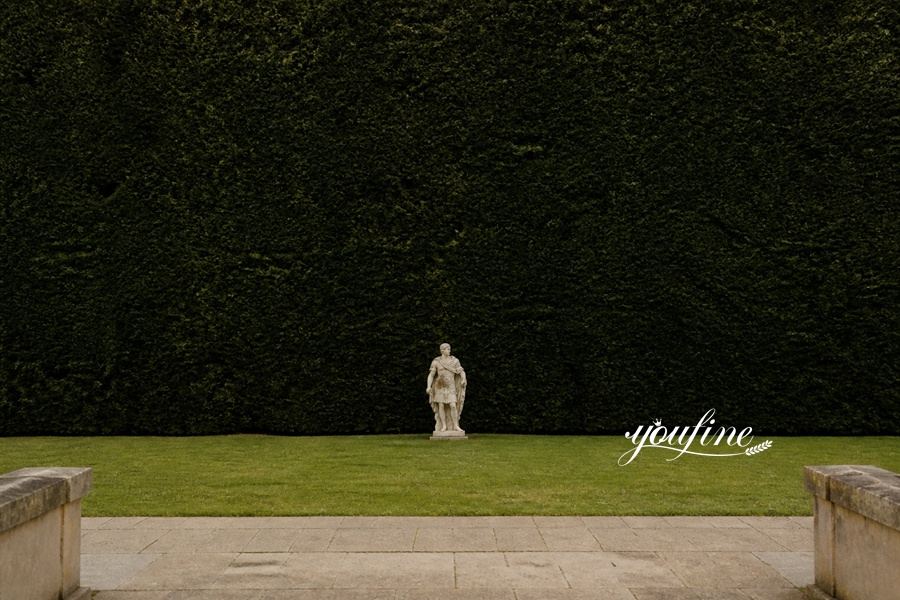 A marble statue of a standing figure in a formal garden with a neatly manicured green hedge background and a stone-paved foreground.
