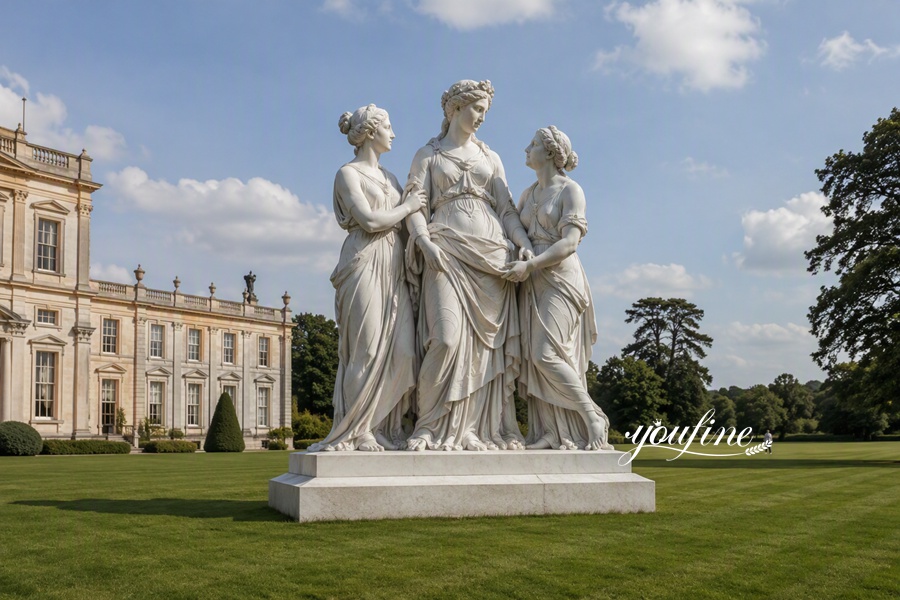 Marble sculpture of The Three Graces featuring three elegantly robed women holding hands, placed in a garden near a historic building.
