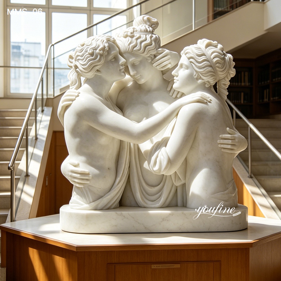 A detailed white marble sculpture depicting the Three Graces in an embrace, placed indoors with natural light and a staircase in the background.