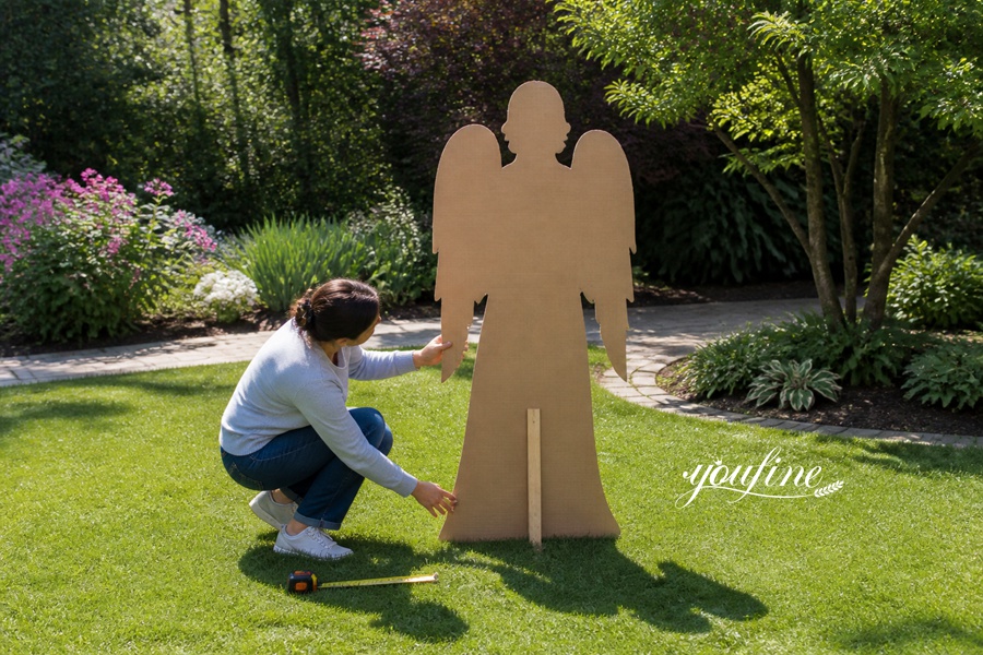 Woman kneeling on a lawn measuring a wooden angel silhouette with a tape measure, surrounded by vibrant garden landscaping.