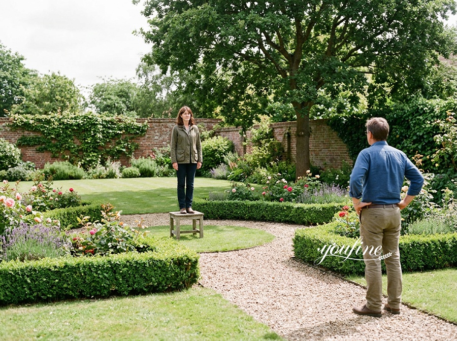 A woman standing on a small wooden stool in the middle of a landscaped garden, surrounded by flowers and greenery, while a man looks on.