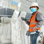 Skilled artisan in an orange vest and mask using a power tool to carve details into a marble statue inside a workshop.