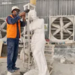 An artisan wearing safety gear polishes a white marble sculpture of a classical female figure in an industrial workshop setting.