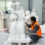 Sculptor wearing an orange vest working on a white marble religious statue featuring a seated figure with two children.