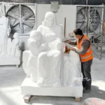 A craftsman wearing an orange vest carves intricate details into a white marble sculpture depicting a seated figure with children, within an industrial workshop.