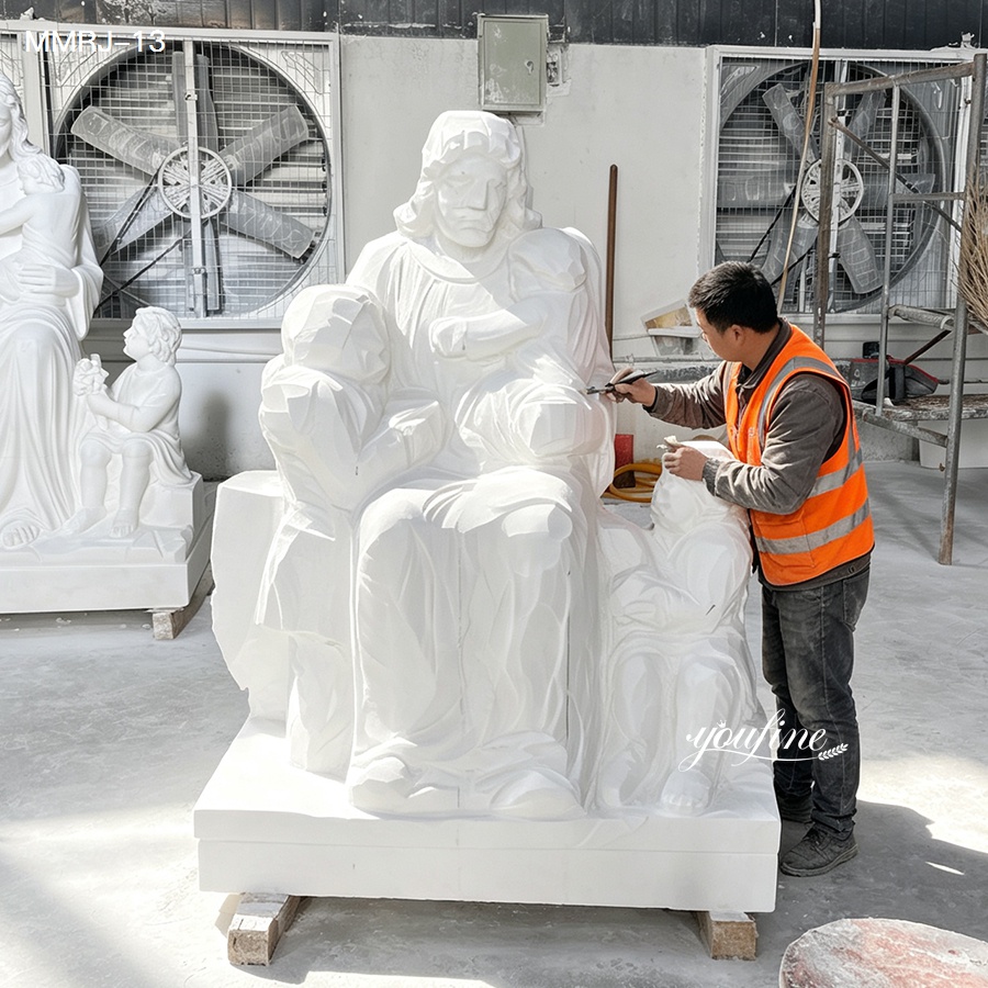 A craftsman wearing an orange vest carves intricate details into a white marble sculpture depicting a seated figure with children, within an industrial workshop.