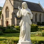 Marble statue of Jesus Christ with hand raised, displayed in a church garden surrounded by vibrant greenery and flowers.