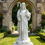 Marble sculpture of Jesus holding a lamb and shepherd's staff placed in a garden with an ornate archway in the background.