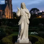 A beautifully carved marble statue of Jesus with hand gestures, placed in a garden near a church at dusk, surrounded by plants.