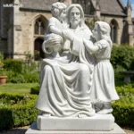 Intricately carved white marble statue of Jesus seated with children, placed in a garden setting near a church building.