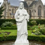 White marble Sacred Heart of Jesus sculpture placed in a garden setting with a church building visible in the background.