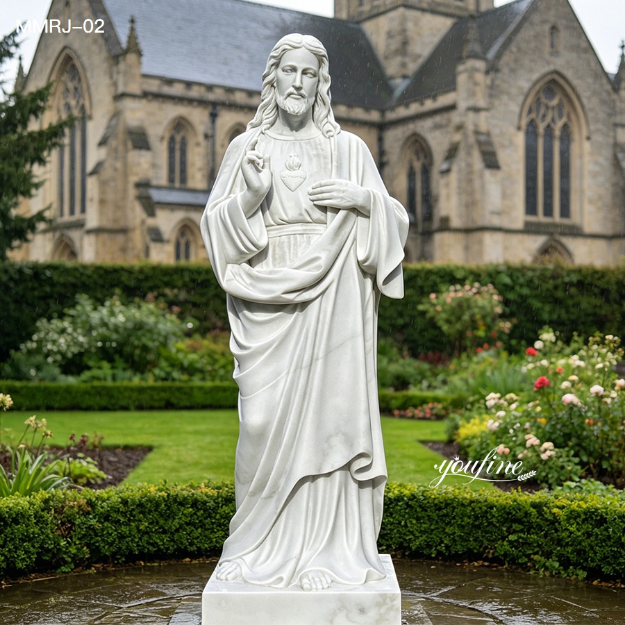 White marble Sacred Heart of Jesus sculpture placed in a garden setting with a church building visible in the background.