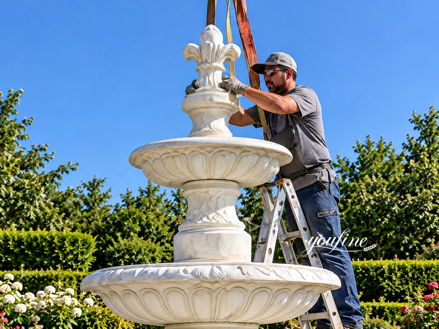 A skilled craftsman installing an ornate white marble fountain with a floral design in a lush green outdoor setting.