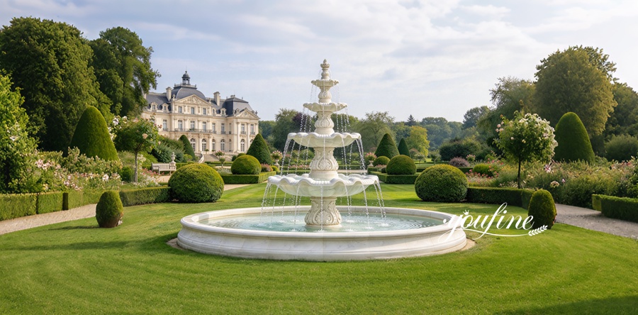 A white marble tiered fountain in a beautifully landscaped garden with manicured greenery and a classical mansion in the background.