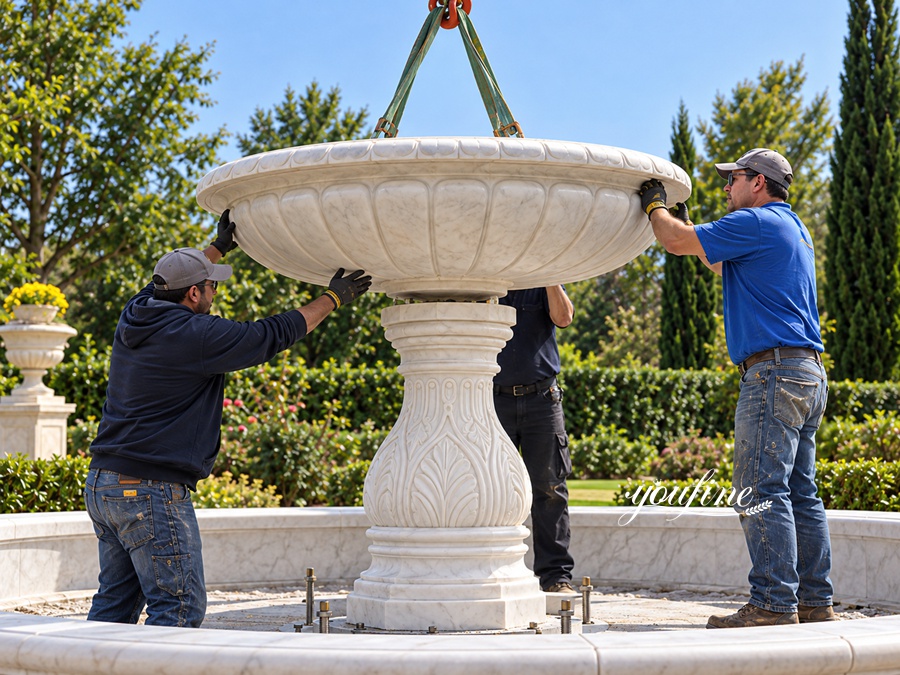 Team of craftsmen installing an ornate marble fountain centerpiece in an outdoor garden setting using crane support.
