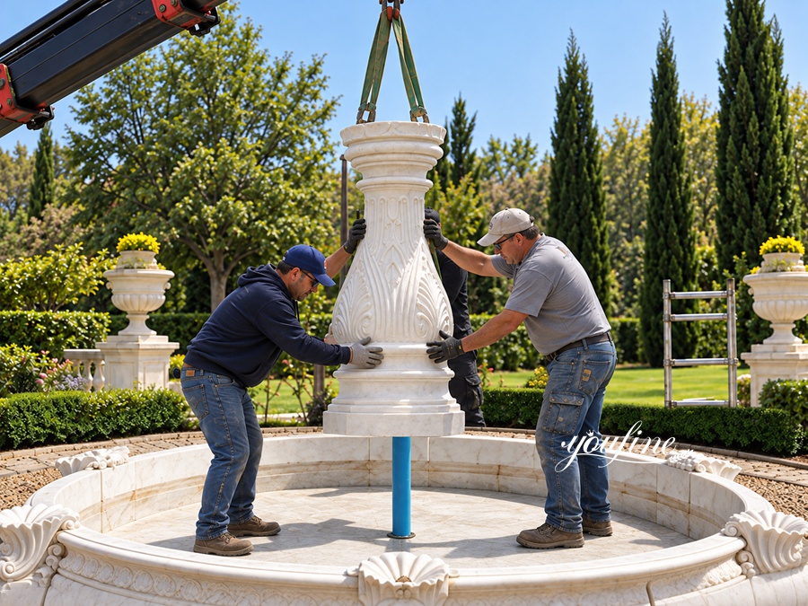 Two workers installing a carved marble fountain centerpiece using a crane in an outdoor landscaped garden.