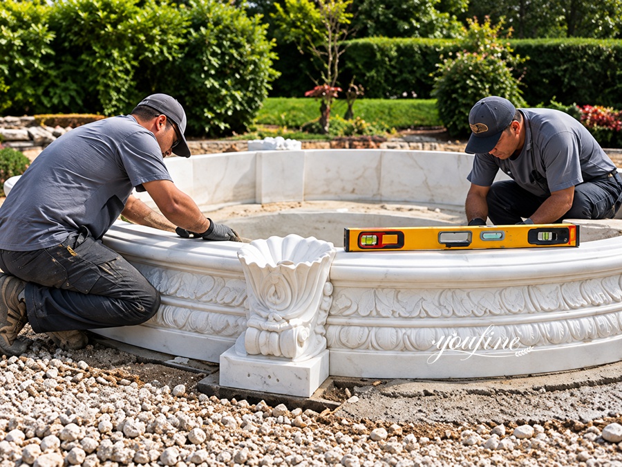 Two craftsmen installing a decorative marble fountain base outdoors, using tools to ensure level alignment in a landscaped setting.